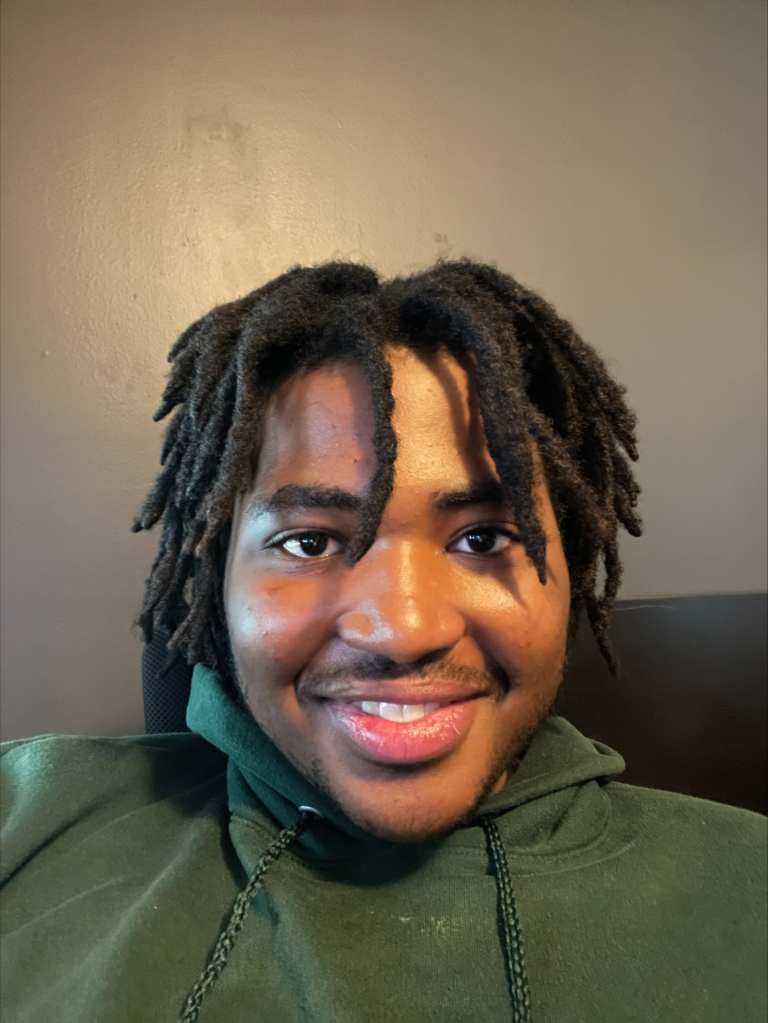 A person with short dreadlocks and a green hoodie is smiling at the camera against a plain brown wall background.