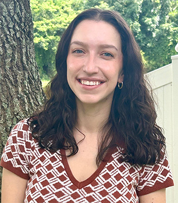 A woman with long, wavy brown hair smiles at the camera while standing outdoors next to a tree, wearing a patterned brown and white shirt. Green foliage and a white fence are in the background.