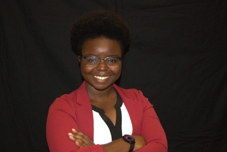 Person wearing glasses, a red blazer, and a white and black shirt stands with arms crossed, smiling in front of a plain black background.