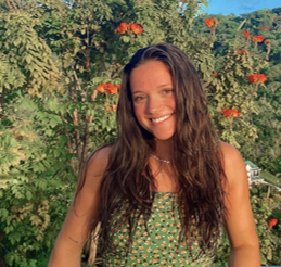 A woman with long brown hair wearing a green patterned dress smiles outdoors with lush greenery and orange flowers in the background.