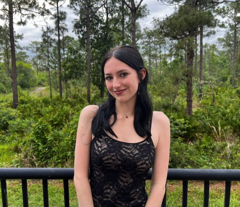 A woman with long black hair in a black lace dress stands in front of a black railing, with a forested area and cloudy sky in the background.