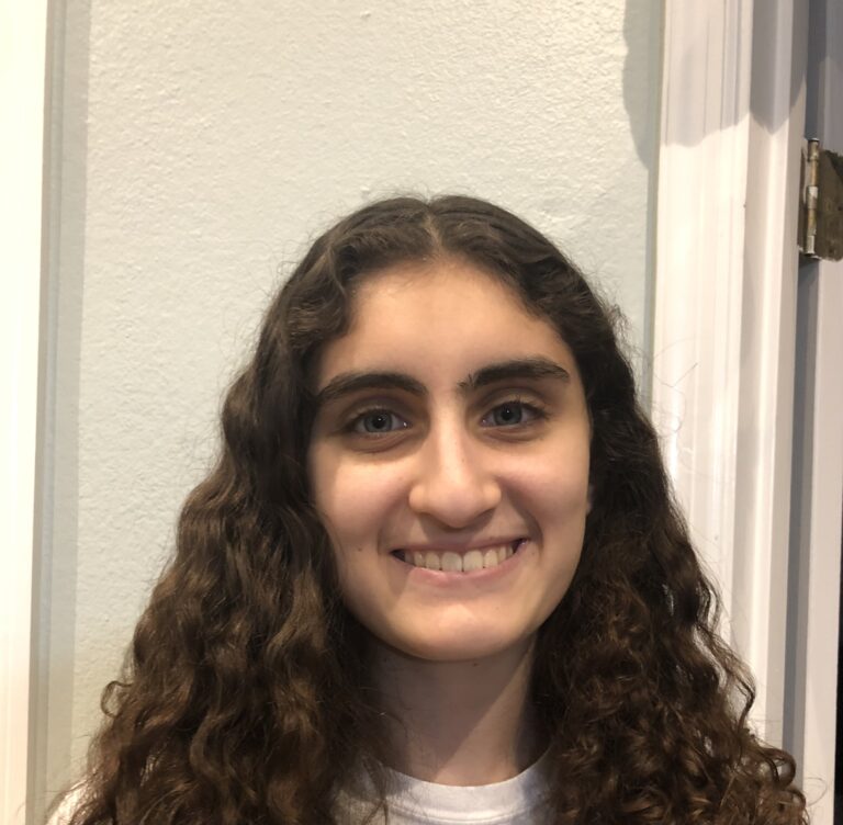 A young person with long, curly brown hair and a white shirt stands indoors, smiling at the camera.