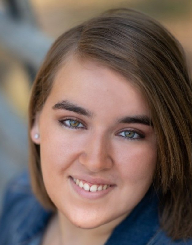 A young woman with straight, light brown hair and hazel eyes smiles at the camera. She is wearing a denim jacket and small stud earrings.