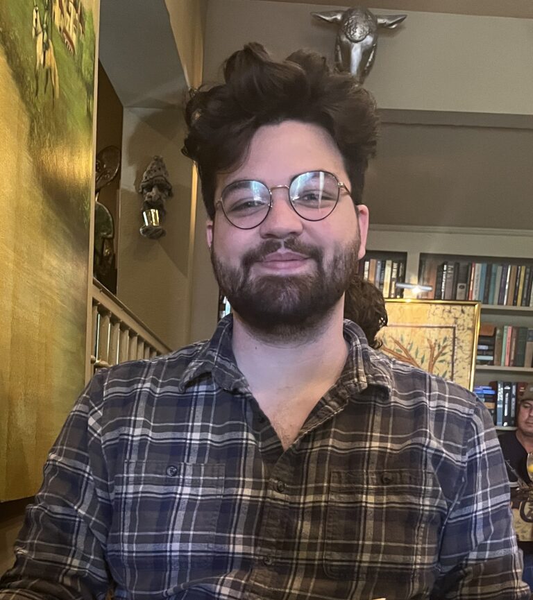 A person with glasses and a beard wearing a plaid shirt sits indoors, with shelves of books and artwork visible in the background.