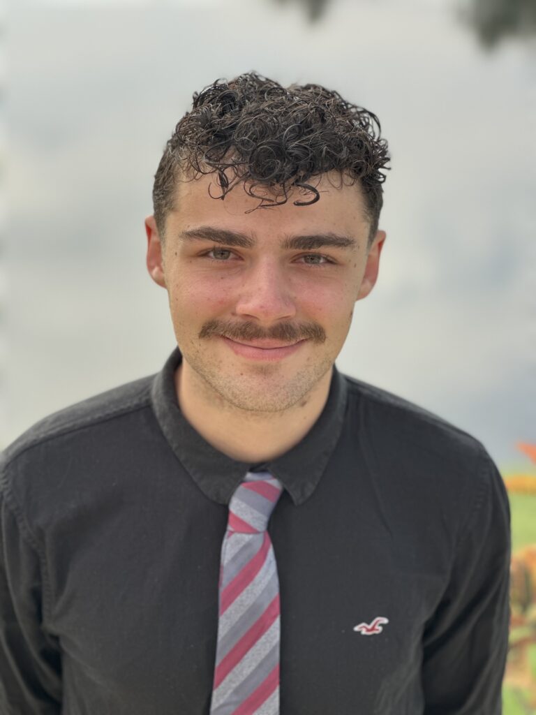 A young man with curly hair and a mustache, wearing a black shirt and a striped tie, stands outdoors in front of a blurred background.