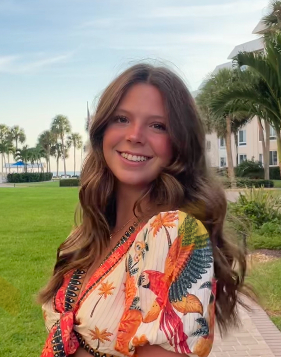 A young woman with long brown hair stands outdoors, smiling, wearing a tropical-patterned top. Palm trees, grass, and buildings are in the background under a blue sky.