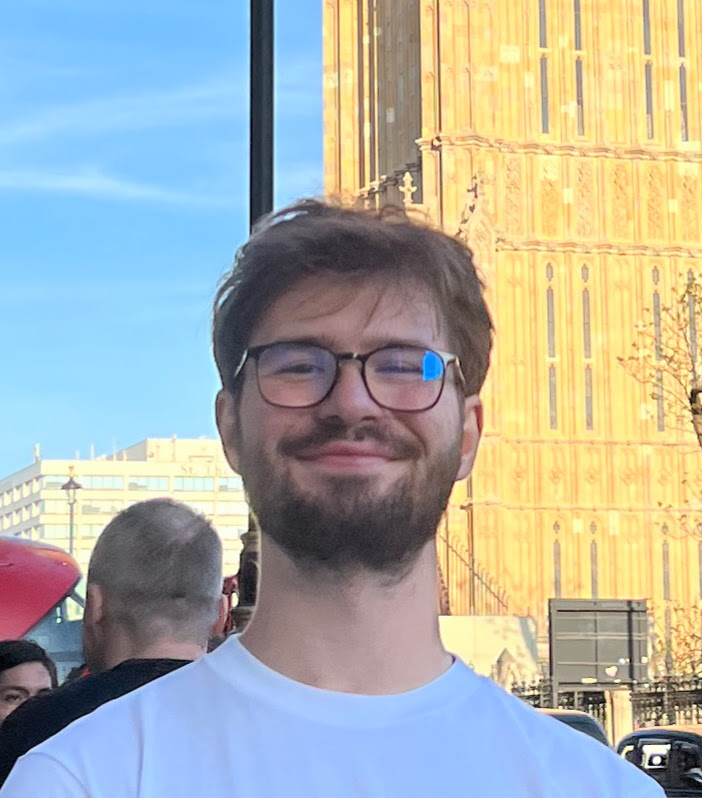 A man with glasses and a beard smiles outdoors in front of a tall building with ornate architecture.