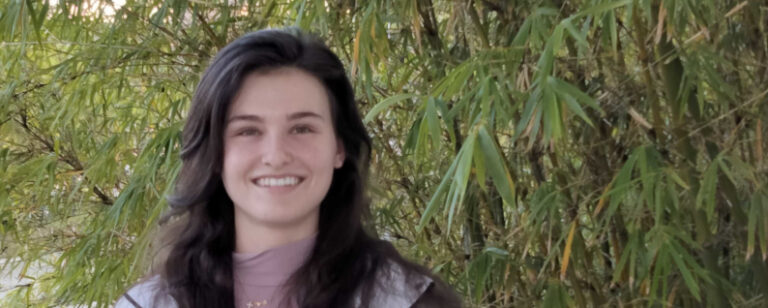 A person with long dark hair and a light purple top smiles while standing in front of green bamboo plants.