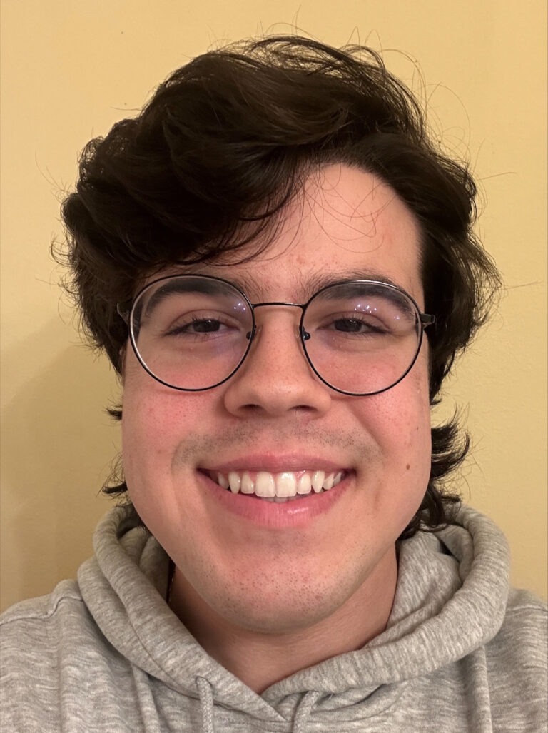 A person with medium-length dark hair, round glasses, and a gray hoodie smiles in front of a plain beige background.