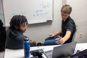 Two people sit at a desk with a laptop, while one points at the screen. A whiteboard with mathematical equations is visible in the background.