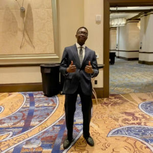 A man in a dark suit stands indoors on patterned carpet, giving two thumbs up, with a conference-style hallway in the background.