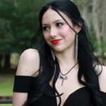 Woman with long black hair, red lipstick, and dangling earrings wearing a black off-shoulder dress and a dragonfly necklace, standing outdoors with trees in the background.