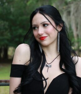 Woman with long black hair, red lipstick, and dangling earrings wearing a black off-shoulder dress and a dragonfly necklace, standing outdoors with trees in the background.