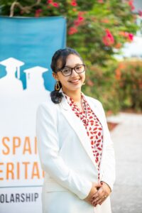 A woman wearing glasses and a white blazer stands outdoors in front of a sign with graduation caps and partial text.