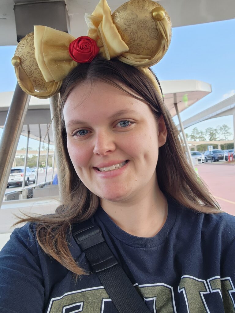 A woman wearing a black shirt and gold Mickey Mouse ears with a red rose smiles at the camera outdoors near parked cars.