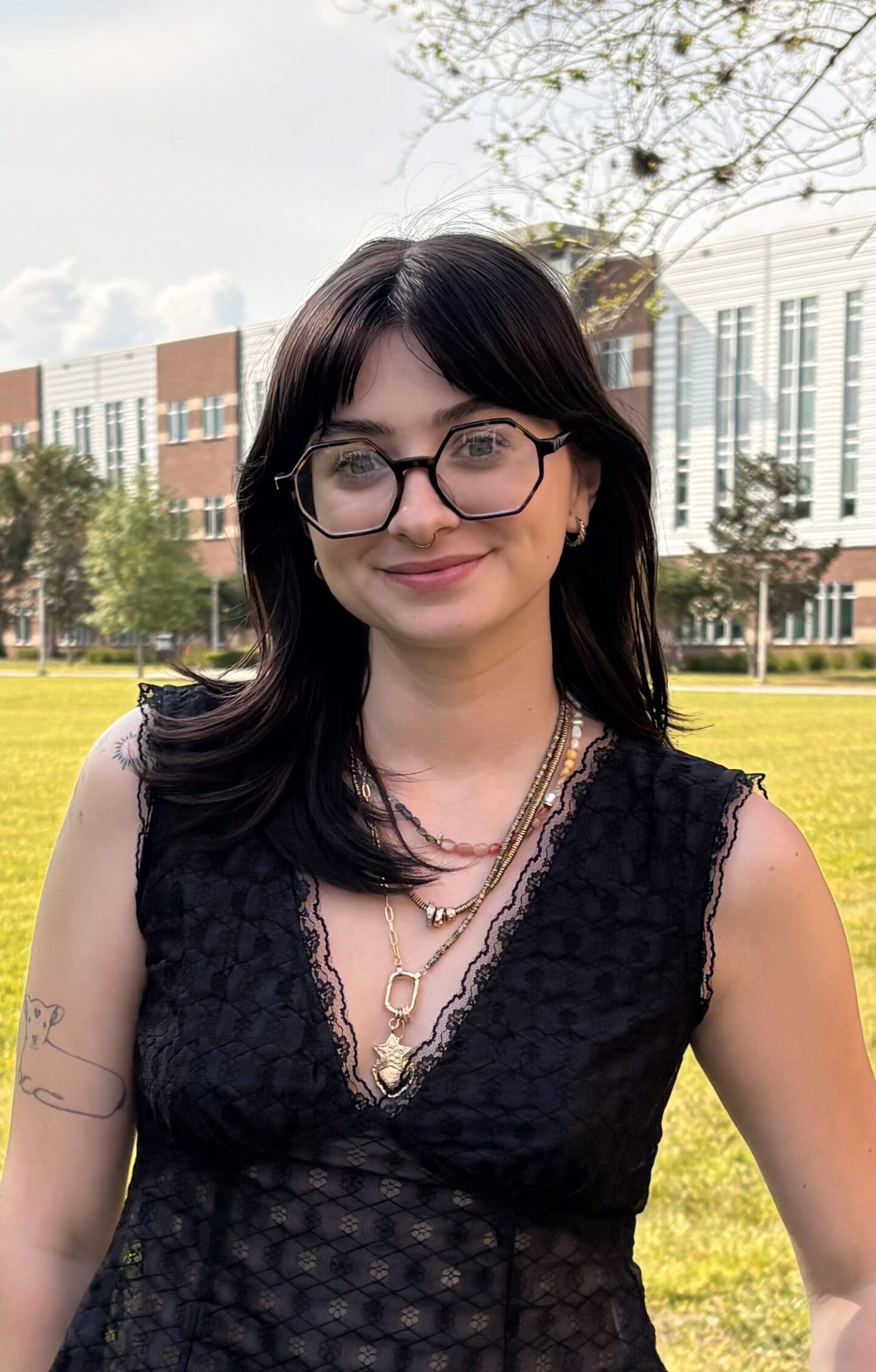 A woman with dark hair and glasses stands outside on a grassy area, wearing a sleeveless black lace top and layered necklaces; buildings and trees are visible in the background.