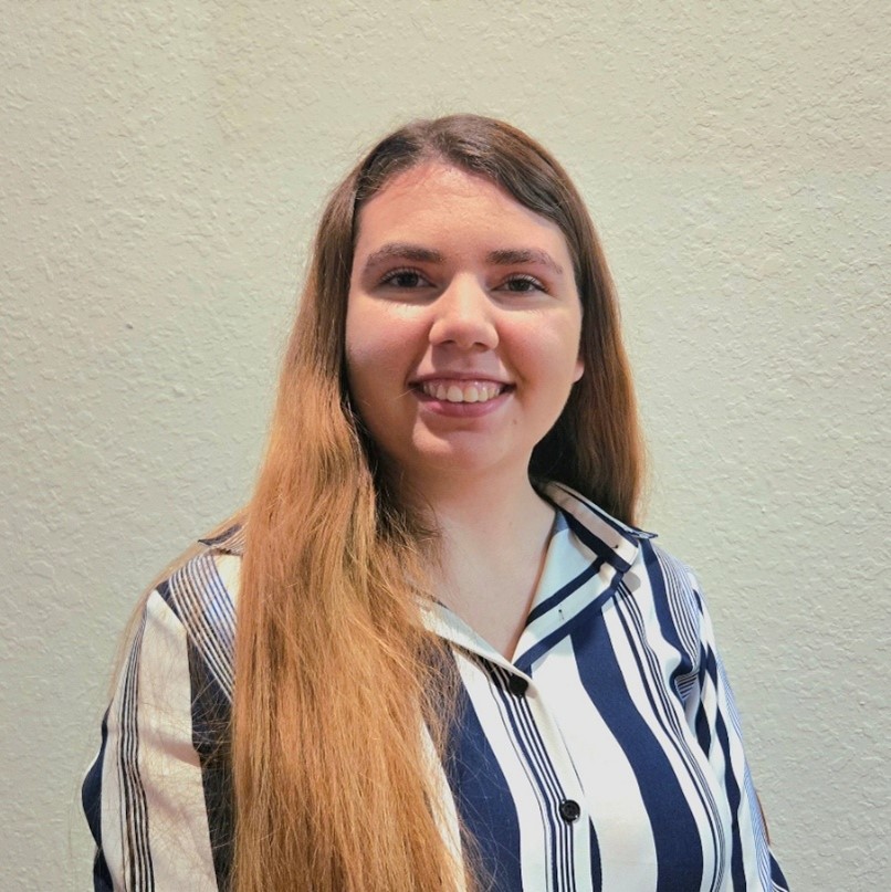 A woman with long brown hair wearing a blue and white vertically striped shirt stands against a light-colored textured wall, smiling at the camera.