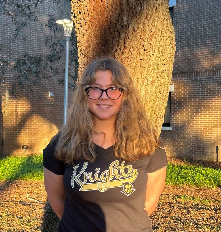 A person with long light brown hair and glasses stands outside in front of a tree, wearing a black UCF Knights t-shirt. Sunlight and tree shadows are visible on the brick wall behind them.