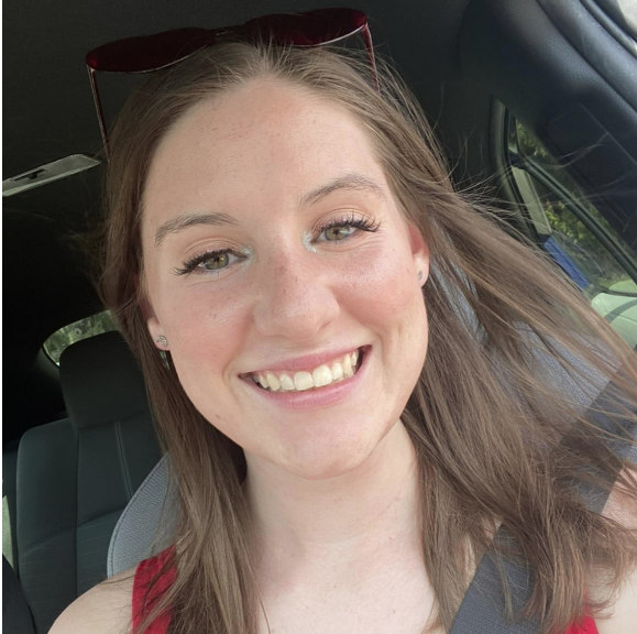 A person with short curly hair, glasses, and a white shirt is smiling outdoors, wearing graduation attire with honor cords. Green grass is visible in the background.