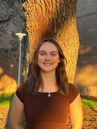 A young woman with brown hair stands in front of a tree, smiling, with sunlight illuminating her face and a brick wall in the background.