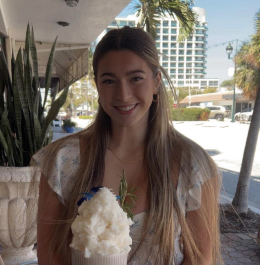 A woman with long hair smiles while holding a dessert topped with whipped cream, sitting outdoors beside a large plant and city buildings in the background.