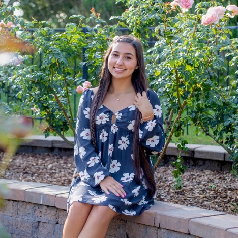 Young woman with long brown hair in a floral dress sits on a low brick wall in a garden with blooming rose bushes, smiling at the camera.