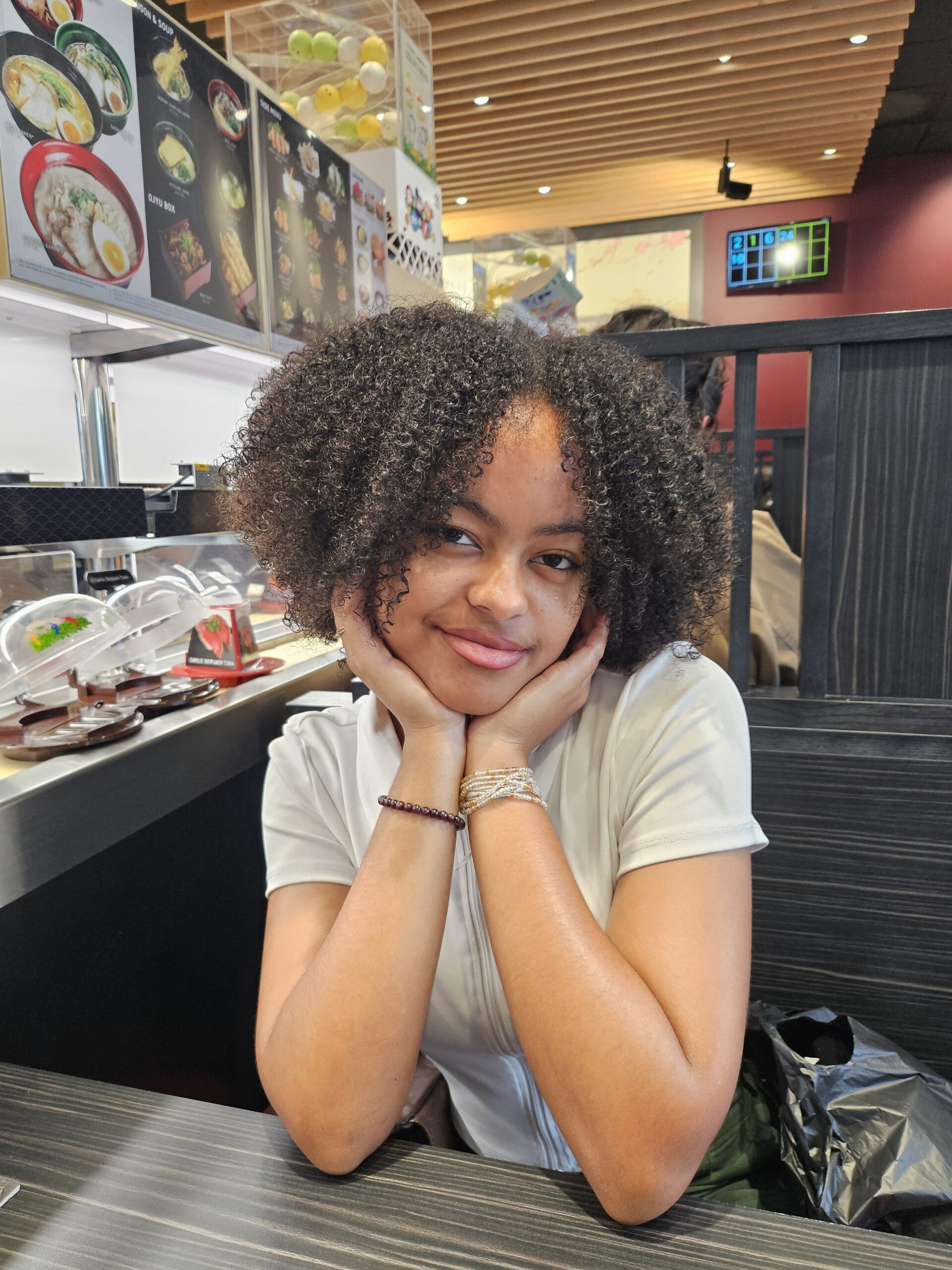 A woman with curly hair sits at a table in a sushi restaurant, resting her chin on her hands and smiling at the camera. Conveyor belt sushi plates are visible in the background.