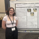 A person stands smiling in front of a scientific research poster presented at a conference, wearing a conference badge and black lanyard.