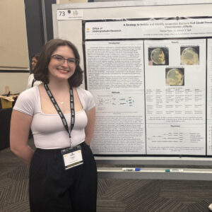 A person stands smiling in front of a scientific research poster presented at a conference, wearing a conference badge and black lanyard.