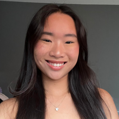 A young woman with long dark hair smiles at the camera, wearing a necklace and a light top, against a plain dark background.