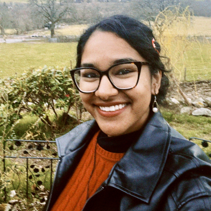 A person wearing glasses, a black jacket, and a red sweater is smiling outdoors in front of greenery and an open field.