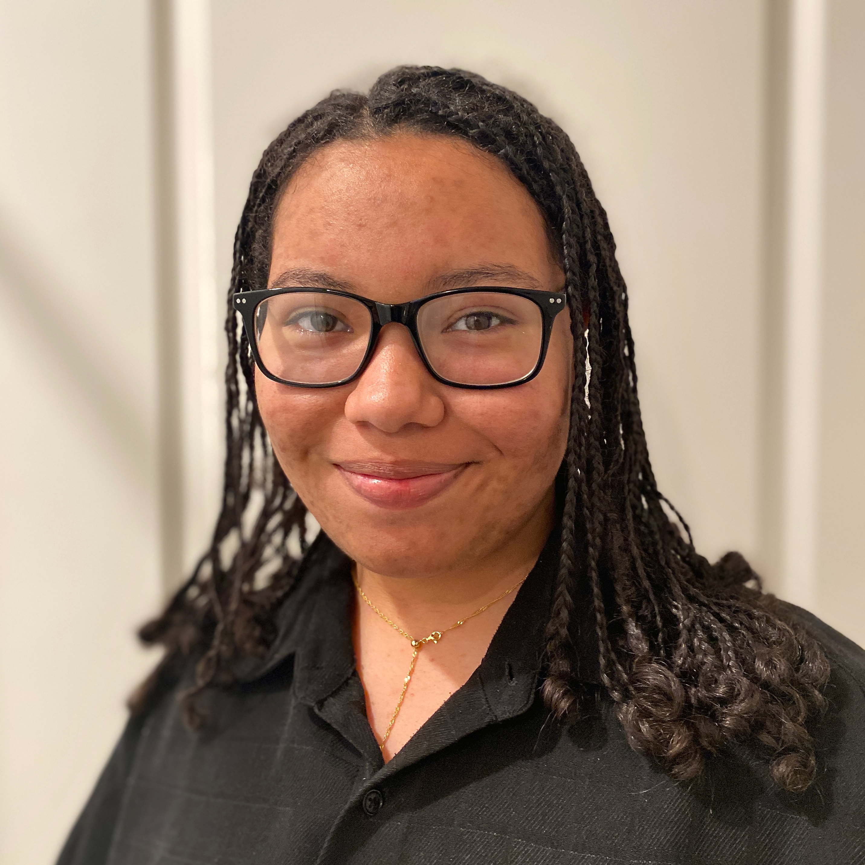 A person with glasses and braided hair smiles slightly, wearing a black shirt and a gold necklace, standing in front of a neutral background.