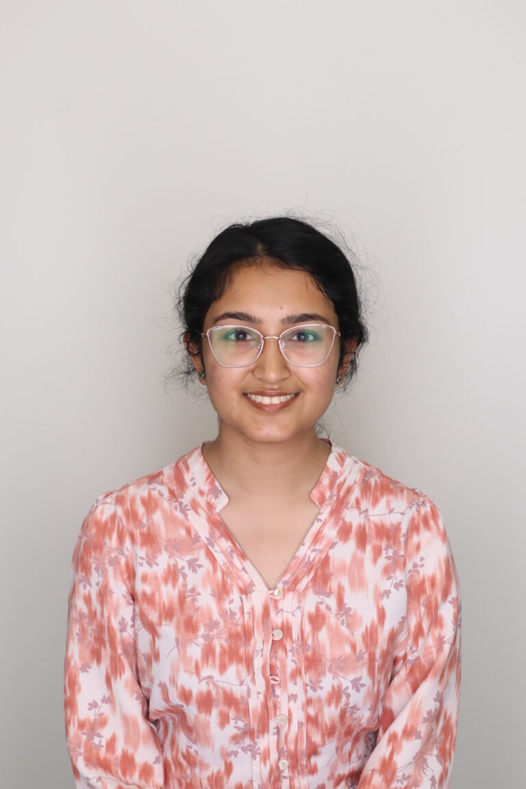 A young woman with glasses and dark hair tied back, wearing a peach and white patterned blouse, sits against a plain light background, smiling at the camera.