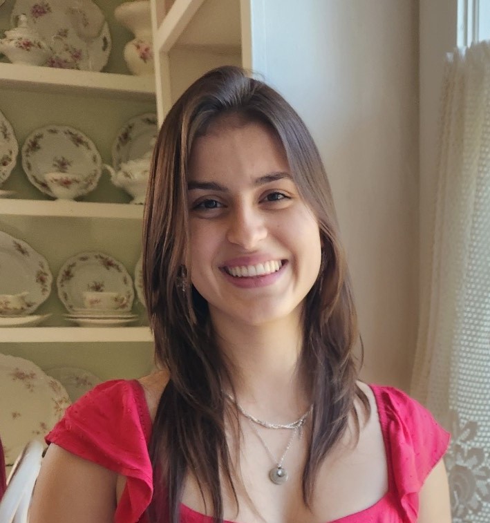A young woman with long brown hair wearing a red top smiles at the camera, standing in front of shelves with decorative plates.