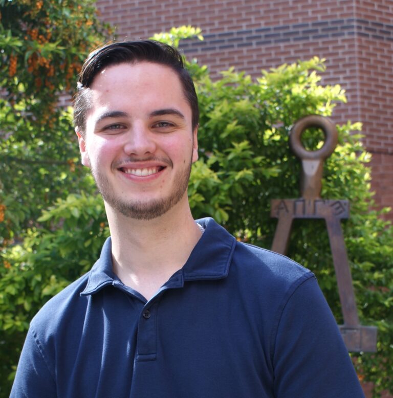 A young man in a navy polo shirt smiles outdoors with green bushes and a metal sculpture in the background.