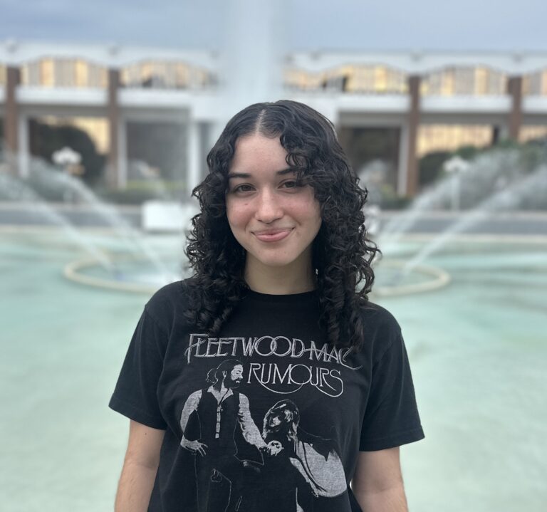 A person with curly hair stands in front of a fountain, wearing a black Fleetwood Mac "Rumours" T-shirt, with a building in the background.