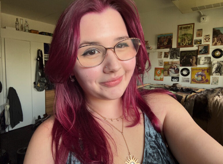 A person with long magenta hair, glasses, and a nose ring smiles in a room decorated with posters and vinyl records.