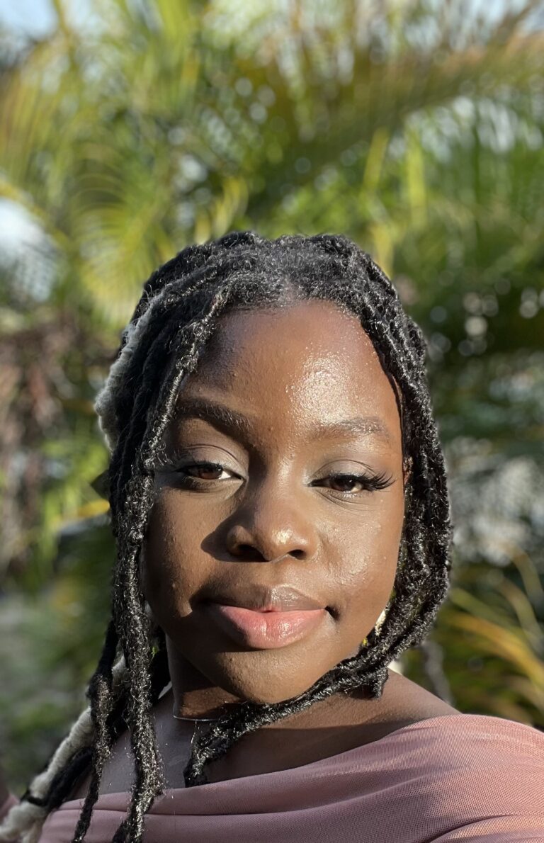 A person with braided hair and a pink top stands outdoors in sunlight, with green palm leaves blurred in the background.