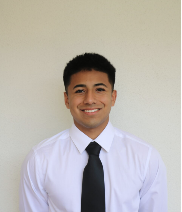 A man with short black hair, wearing a white dress shirt and black tie, stands against a plain light-colored background, smiling at the camera.