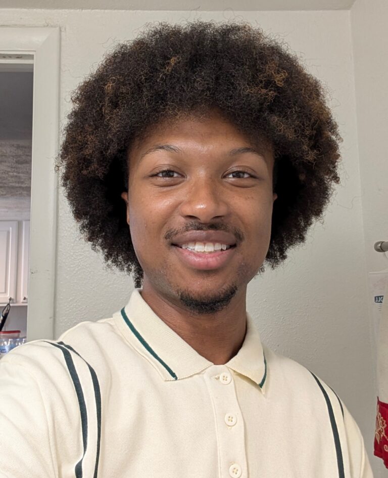A person with short facial hair and a curly afro hairstyle smiles at the camera, wearing a cream collared shirt with green stripes, standing indoors.