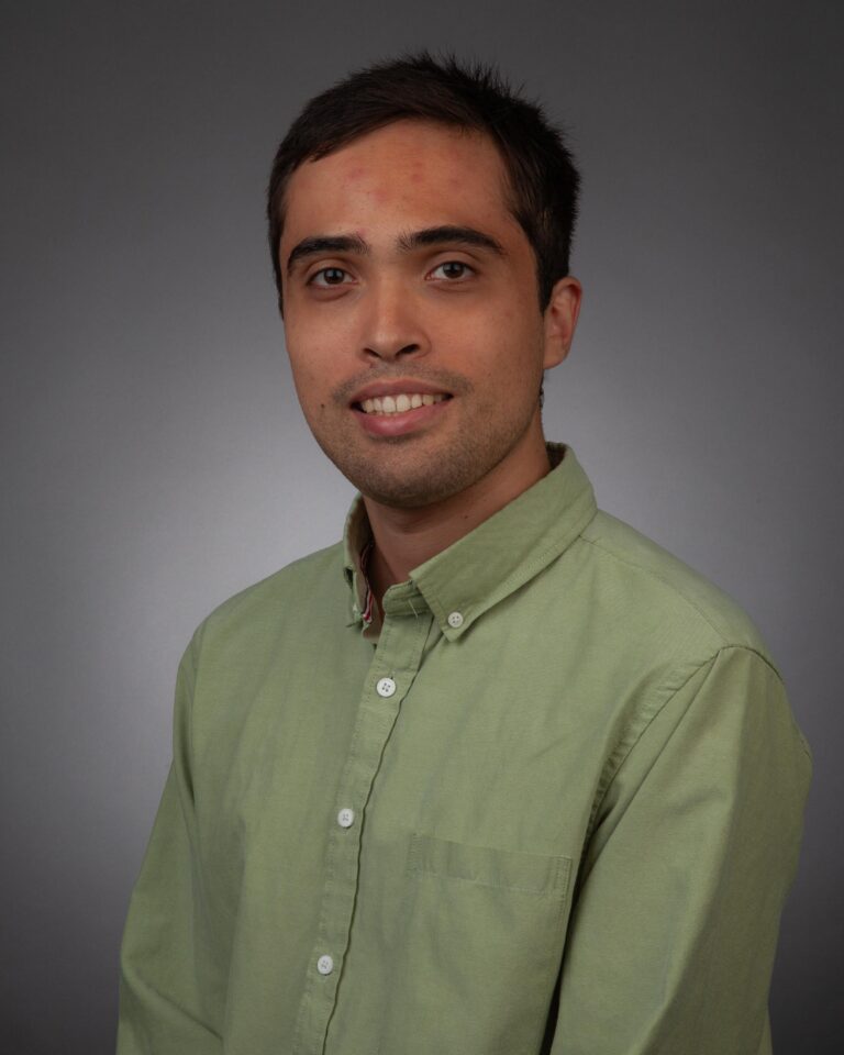 A man with short dark hair wearing a light green button-up shirt poses in front of a plain gray background.