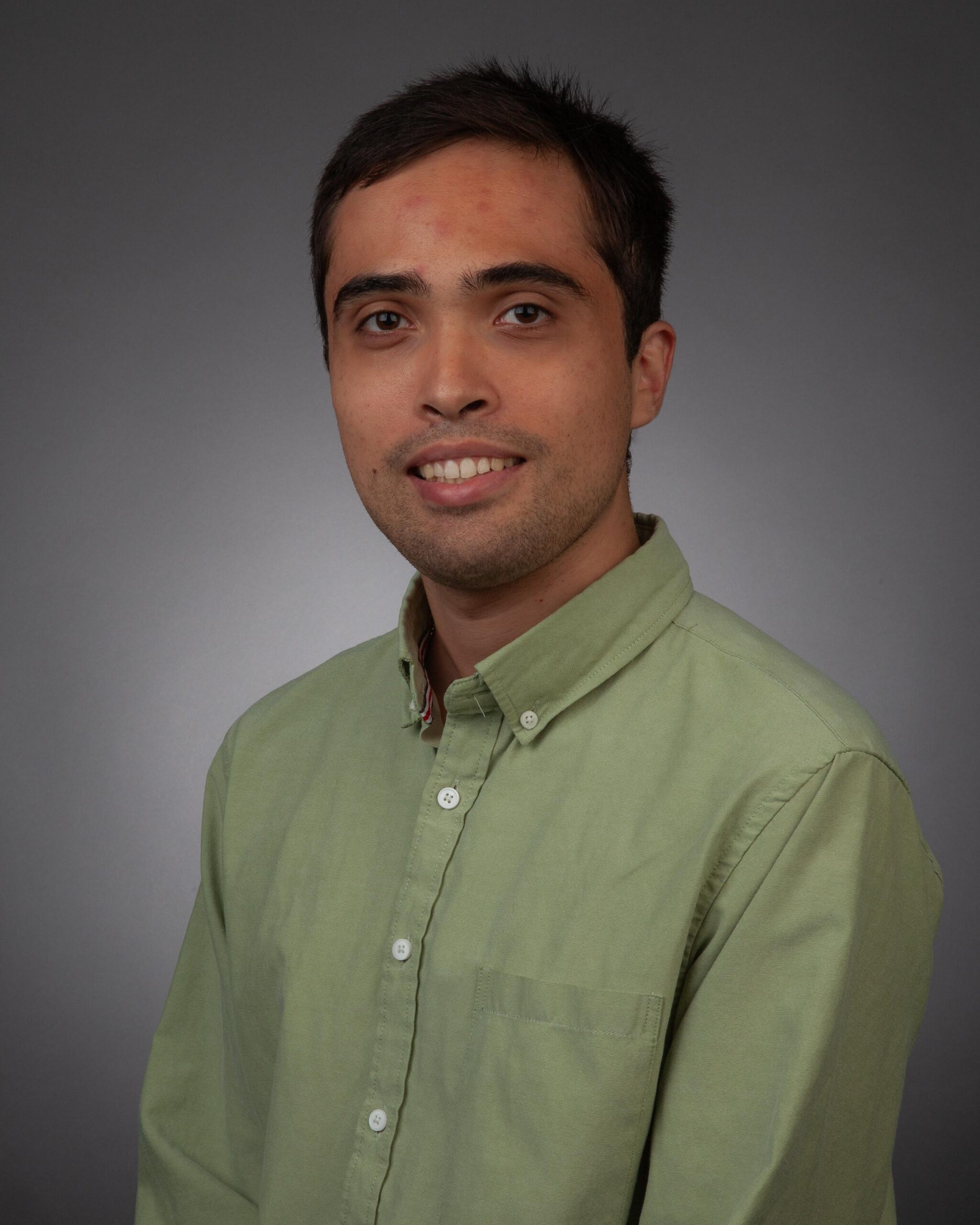 A man with short dark hair wearing a light green button-up shirt poses in front of a plain gray background.