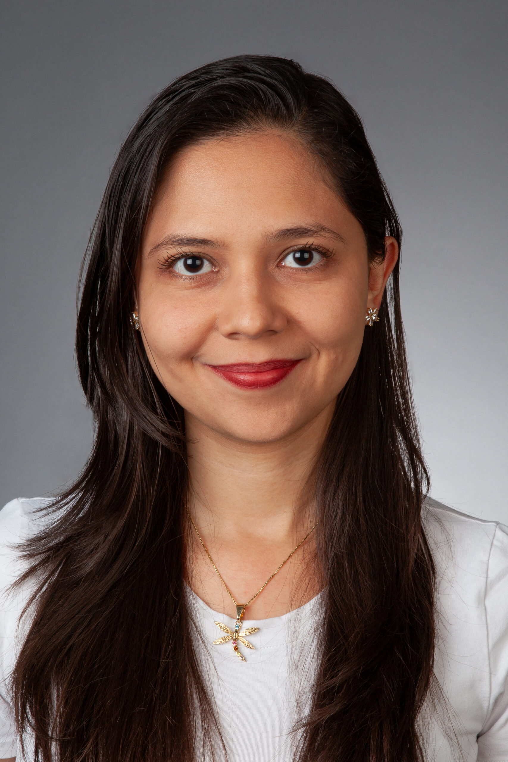 A woman with long brown hair, wearing a white top, gold necklace, and red lipstick, smiles at the camera against a plain gray background.