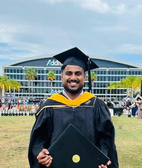 A graduate in cap and gown smiles while holding his diploma folder outside a large building with "Addition" on its front.