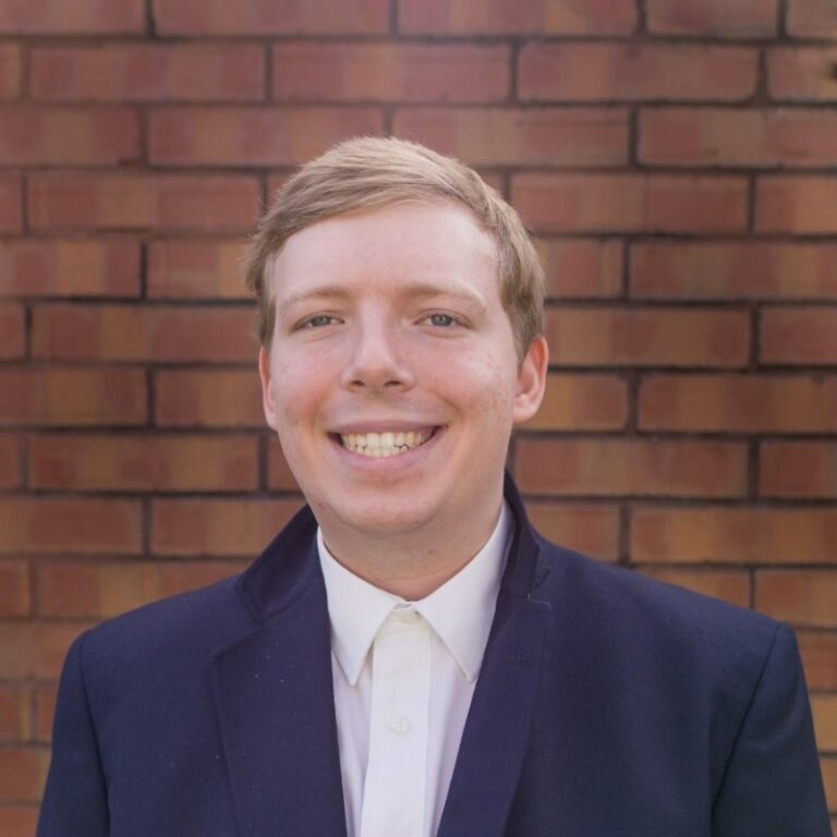 A man in a dark blazer and white shirt smiles at the camera, standing in front of a red brick wall.