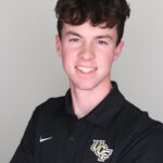 Young man with short brown hair wearing a black UCF polo shirt, smiling and standing against a plain light background.