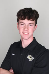 Young man with short brown hair wearing a black UCF polo shirt, smiling and standing against a plain light background.