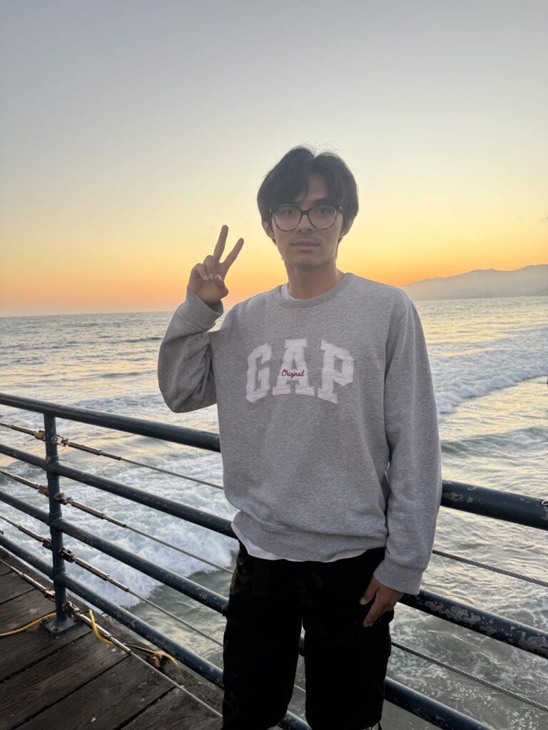 A person wearing glasses and a gray GAP sweatshirt stands on a pier, making a peace sign with one hand, with the ocean and sunset in the background.