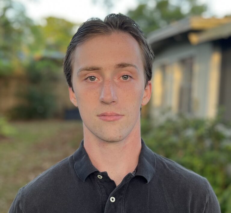 A young man with short brown hair and fair skin stands outside, facing the camera, wearing a black collared shirt. The background is blurred, showing greenery and part of a house.