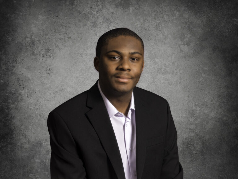 A man wearing a black suit jacket and white shirt poses for a formal portrait in front of a gray textured background.